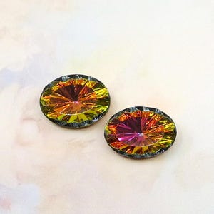 May include: Two iridescent, oval-shaped gemstones with a faceted design. The gems display a spectrum of colors, including yellow, orange, and pink, with a dark gray border. The gemstones are set against a soft, light pink background.