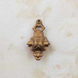 Puede incluir: Un colgante de metal dorado con detalles intrincados y un diseño en forma de rollo. El colgante tiene un bucle en la parte superior para sujetarlo a una cadena o cordón.