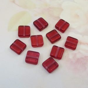May include: A collection of ten square, red glass beads with a white stripe across the center. The beads are arranged on a light-colored surface, showcasing their translucent quality and reflective surfaces. These beads are ideal for jewelry making.