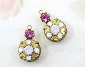 May include: Two gold-toned metal charms, each featuring a circular base set with a central pale pink crystal surrounded by four small white crystals, topped with a single vibrant pink crystal and a small circular loop for attachment.