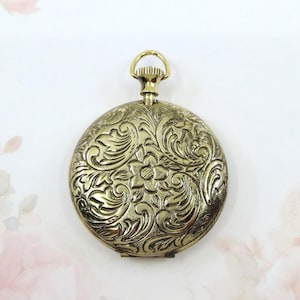 May include: Antique gold-colored pocket watch case with intricate floral and scrollwork engraving. The round case features a detailed design with a central flower and swirling patterns. A small loop is at the top for attaching a chain.