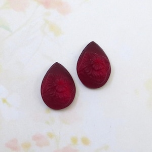 May include: Two dark red teardrop-shaped cabochons with a carved floral design. The cabochons are displayed on a white and light pink patterned background. Suitable for jewellery making, these cabochons are a rich, deep red colour.