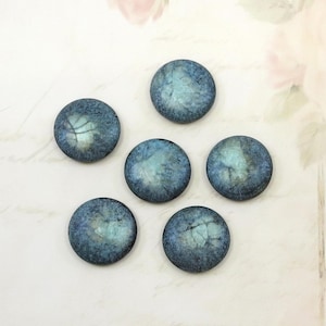 May include: Six circular, blue-green cabochons with a marbled effect. Each cabochon features a dark blue outer edge and a lighter, iridescent centre. They are arranged on a white surface, with soft pink floral accents in the background.