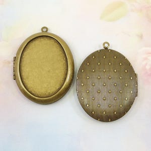 May include: Two antique bronze oval lockets. One locket is open, revealing a blank interior. The other locket is closed, with a textured, patterned design on the exterior. Both lockets have a small loop at the top for a chain.
