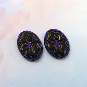 Puede incluir: Dos botones ovalados negros con un diseño de estrella. Los botones tienen un brillo metálico con detalles en azul, dorado y morado. El centro de la estrella es de color azul vibrante.
