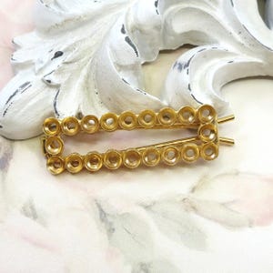 May include: A gold-coloured rectangular hair barrette with a series of small, circular openings along its edges. The barrette has two straight pins on the back for securing it in the hair. The barrette is a hair accessory.