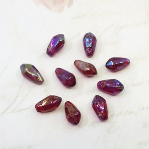 May include: A collection of iridescent, teardrop-shaped beads in shades of purple, red, and gold. Each bead has a hole for stringing. The beads are scattered on a white surface, ideal for jewelry making.