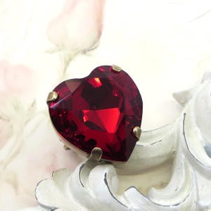 Może przedstawiać: Fasetowany, w kształcie serca, głęboko czerwony kamień szlachetny ze złotymi pazurami. Kamień szlachetny jest osadzony na białym, ozdobnym tle z miękkim kwiatowym wzorem.