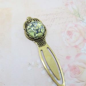 May include: A vintage-style brass bookmark with an oval, green and blue glass cabochon. The bookmark features ornate detailing around the cabochon and along the handle. The background is a soft pink with floral accents.