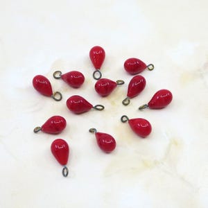 May include: A collection of glossy, teardrop-shaped red beads with small, antique brass-colored loops. These beads are scattered across a light-colored surface, showcasing their vibrant color and reflective quality. Ideal for jewelry making.