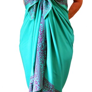 Teal green sarong with a purple floral border that goes all the way around on all four sides. It is tied as a dress with a bow at the breast line. The border design flows down the front, across the breasts, and hems the bottom at the knee
