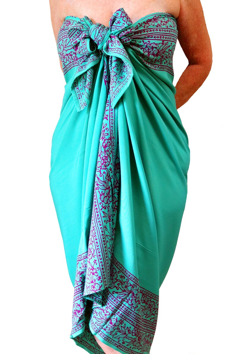 Teal green sarong with a purple floral border that goes all the way around on all four sides. It is tied as a dress with a bow at the breast line. The border design flows down the front, across the breasts, and hems the bottom at the knee