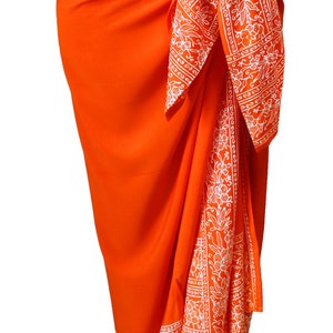 Deep solid orange sarong wrapped as a skirt and tied at the waist. It has a 6 inch white floral border that goes all the way around on all four sides. The border wraps around the hips, drapes down the front and lines the bottom at the ankles.