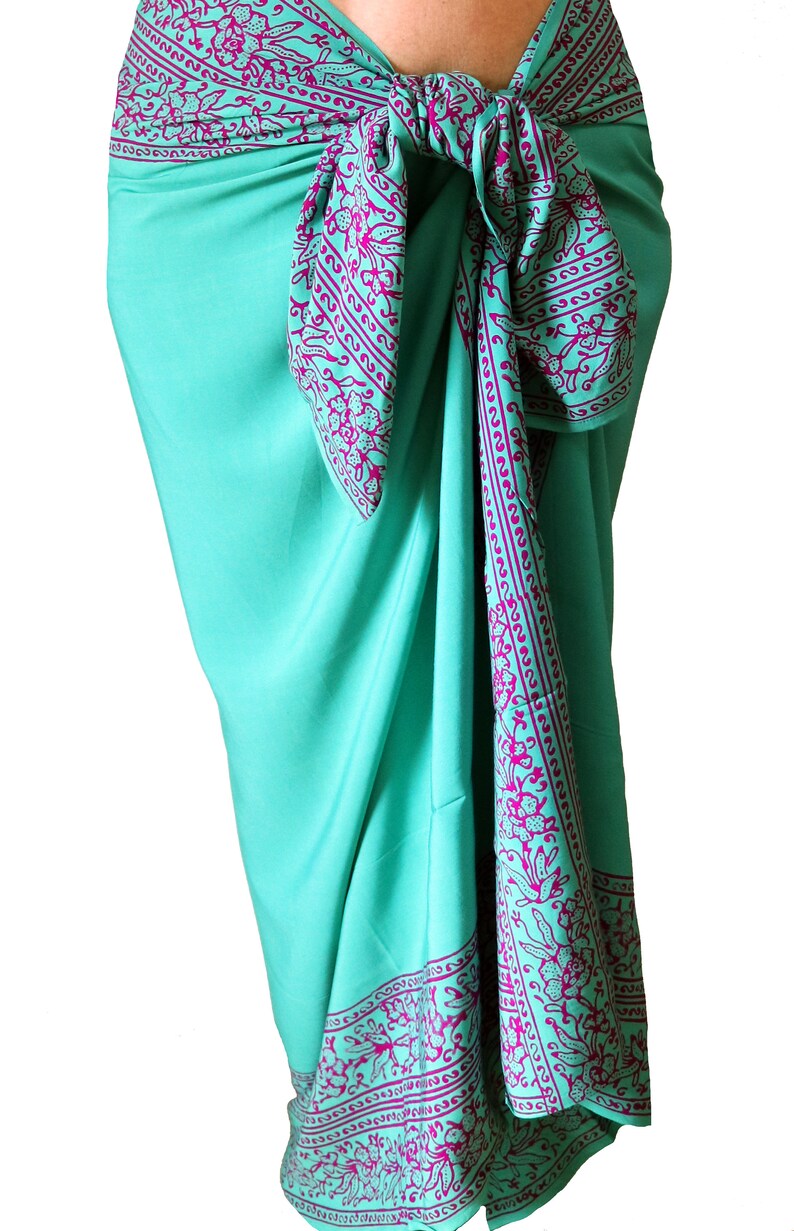 Teal green sarong wrapped as a skirt and tied at the waist. It has a 6 inch purple floral border that goes all the way around on all four sides. The purple border wraps around the hips, drapes down the front and lines the bottom at the ankles.