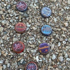 May include: Seven bottle caps from Nuka Cola, a fictional beverage brand from the Fallout video game series. The caps are various colors, including red, blue, and white, and feature different designs, including the words "Nuka Cola", "Sunset Sarsaparilla", "Victory", and "Quantum".