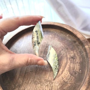 May include: Two silver-colored, leaf-shaped earrings with a textured surface. The earrings are held against a wooden background. The earrings have a reflective quality, catching the light and reflecting the surrounding environment.
