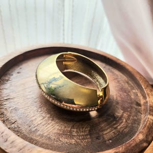May include: A gold-toned hinged bangle bracelet with a row of small, clear crystals along one edge. The bracelet is resting on a round, wooden tray with a dark finish. The bracelet has a smooth, polished surface.