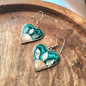 May include: A pair of heart-shaped earrings with a silver-colored metal border. The earrings feature a teal-colored enamel background with inlaid iridescent shell and white leaf-shaped accents. The earrings are displayed on a wooden surface.