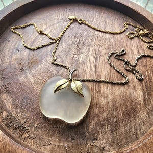 May include: A vintage necklace featuring a frosted glass apple pendant with gold-toned leaves. The pendant hangs from a delicate gold-toned chain. The necklace is displayed on a wooden tray with a rustic finish.