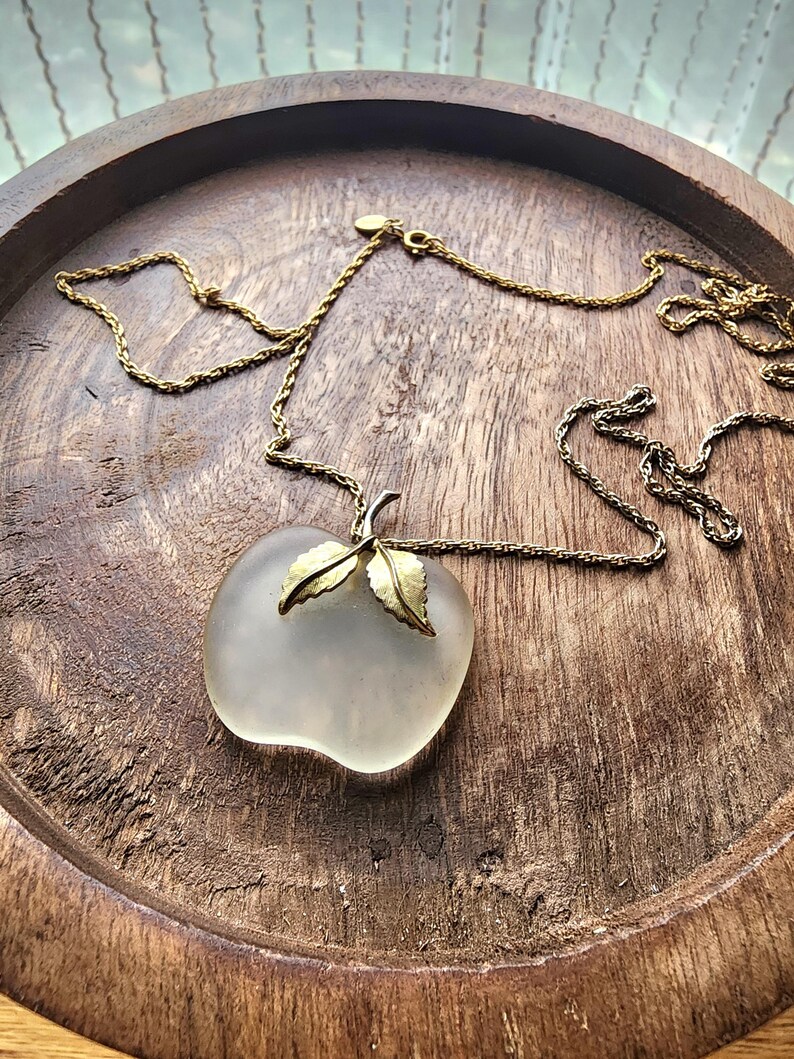 May include: A vintage necklace featuring a frosted glass apple pendant with gold-toned leaves. The pendant hangs from a delicate gold-toned chain. The necklace is displayed on a wooden tray with a rustic finish.
