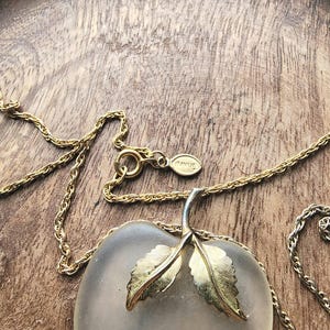May include: A gold-toned necklace with a frosted glass apple pendant. The apple pendant has gold-colored leaves and a silver stem. The chain is delicate and the clasp has an "AVON" tag. The necklace rests on a wooden surface.