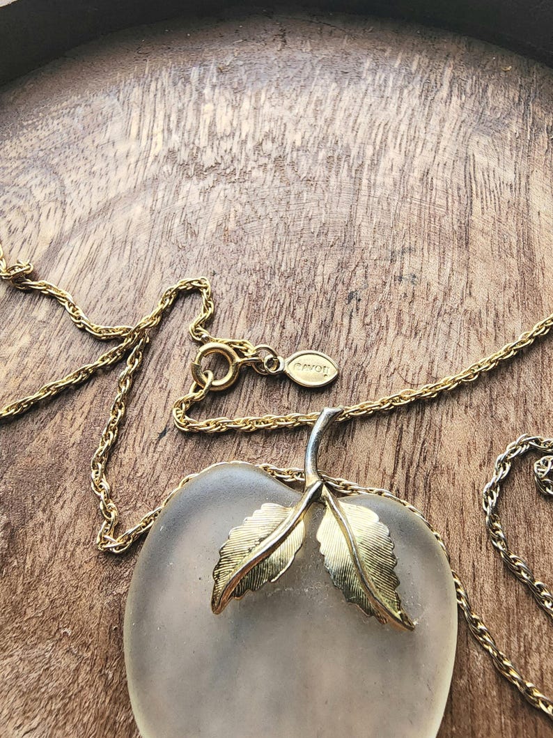 May include: A gold-toned necklace with a frosted glass apple pendant. The apple pendant has gold-colored leaves and a silver stem. The chain is delicate and the clasp has an "AVON" tag. The necklace rests on a wooden surface.