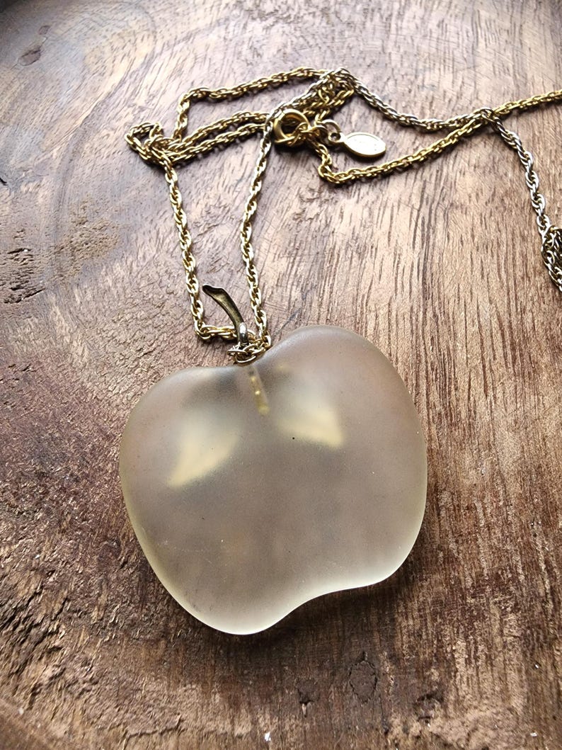 May include: A gold-toned necklace with a frosted glass apple pendant. The apple pendant is a light, translucent color and has a stem detail. The necklace chain is delicate and has a lobster clasp.