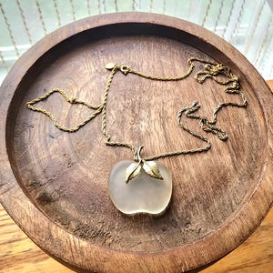 May include: A gold-toned necklace with a frosted glass apple pendant. The apple pendant has gold-colored leaves and stem. The necklace is displayed on a round, wooden tray.