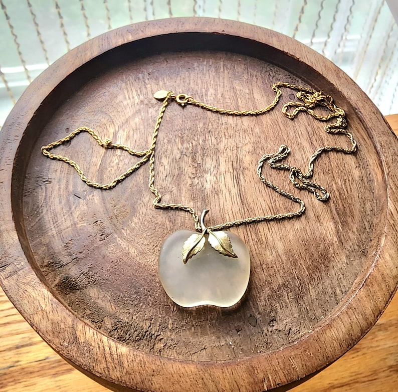 May include: A gold-toned necklace with a frosted glass apple pendant. The apple pendant has gold-colored leaves and stem. The necklace is displayed on a round, wooden tray.