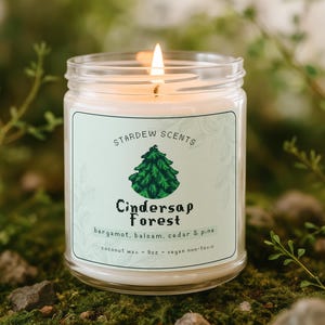 May include: A lit glass jar candle with a label that reads "Cindersap Forest" with a green tree graphic. The candle is made with coconut wax and has a 255g capacity. The scent includes bergamot, balsam, cedar, and pine.