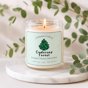 May include: A lit glass jar candle with a label that reads "Stardew Scents Cindersap Forest" with a green tree graphic. The candle is on a white marble surface, surrounded by green leaves. The candle is 255g and made of coconut wax.