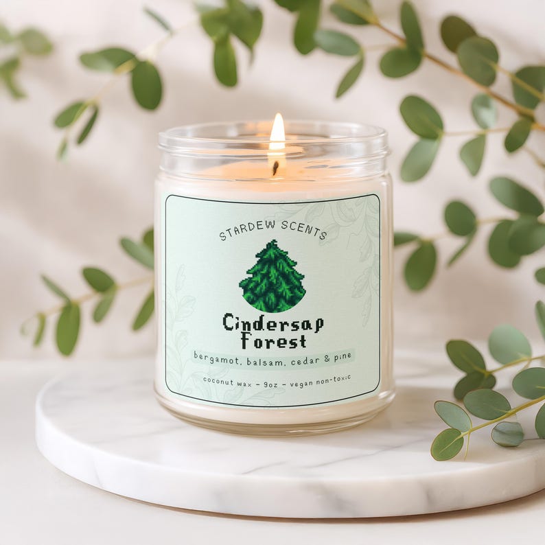 May include: A lit glass jar candle with a label that reads "Stardew Scents Cindersap Forest" with a green tree graphic. The candle is on a white marble surface, surrounded by green leaves. The candle is 255g and made of coconut wax.