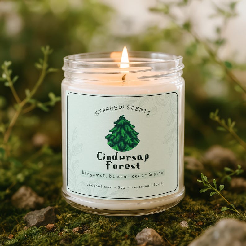 May include: A lit glass jar candle with a label that reads "Cindersap Forest" with a green tree graphic. The candle is made with coconut wax and has a 255g capacity. The scent includes bergamot, balsam, cedar, and pine.