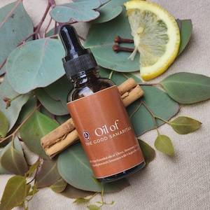 Puede incluir: Un frasco cuentagotas de vidrio ámbar oscuro con tapón negro y etiqueta marrón con el texto Oil of THE GOOD SAMARITAN. La etiqueta enumera ingredientes como clavo, romero, eucalipto y canela. El frasco está rodeado de hojas de eucalipto.
