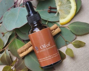 May include: A dark amber glass dropper bottle with a black cap and a brown label reading Oil of THE GOOD SAMARITAN. The label lists ingredients including clove, rosemary, eucalyptus, and cinnamon. The bottle is surrounded by eucalyptus leaves.