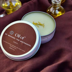 Puede incluir: Una lata de plata con una etiqueta marrón que dice "Aceite de San Miguel Arcángel Bálsamo" y enumera los ingredientes como "Lavanda, Geranio, Caléndula, Aceite de Coco Extra Virgen y Cera de Abeja". La lata contiene 60 ml de bálsamo verde claro.