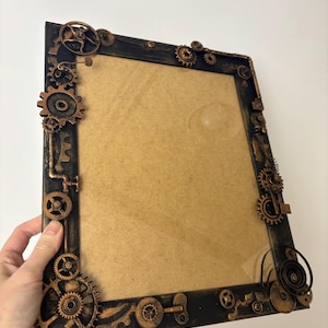 May include: A black picture frame with a steampunk design. The frame is decorated with gears, pipes, and other metal accents. The frame is made of wood and has a distressed finish.
