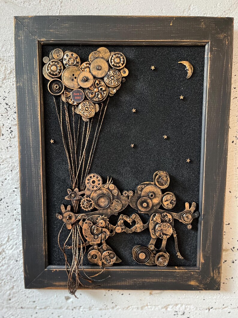 May include: A framed artwork featuring a steampunk-style Mickey Mouse figure made of gears and other metal pieces. The figure is holding a heart-shaped balloon made of buttons and gears, and is flying against a black background with gold stars and a crescent moon.