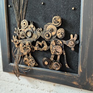 May include: A framed artwork featuring a steampunk-inspired design with gears, cogs, and other metal elements arranged in a whimsical, abstract shape. The background is a dark black with gold glitter stars.
