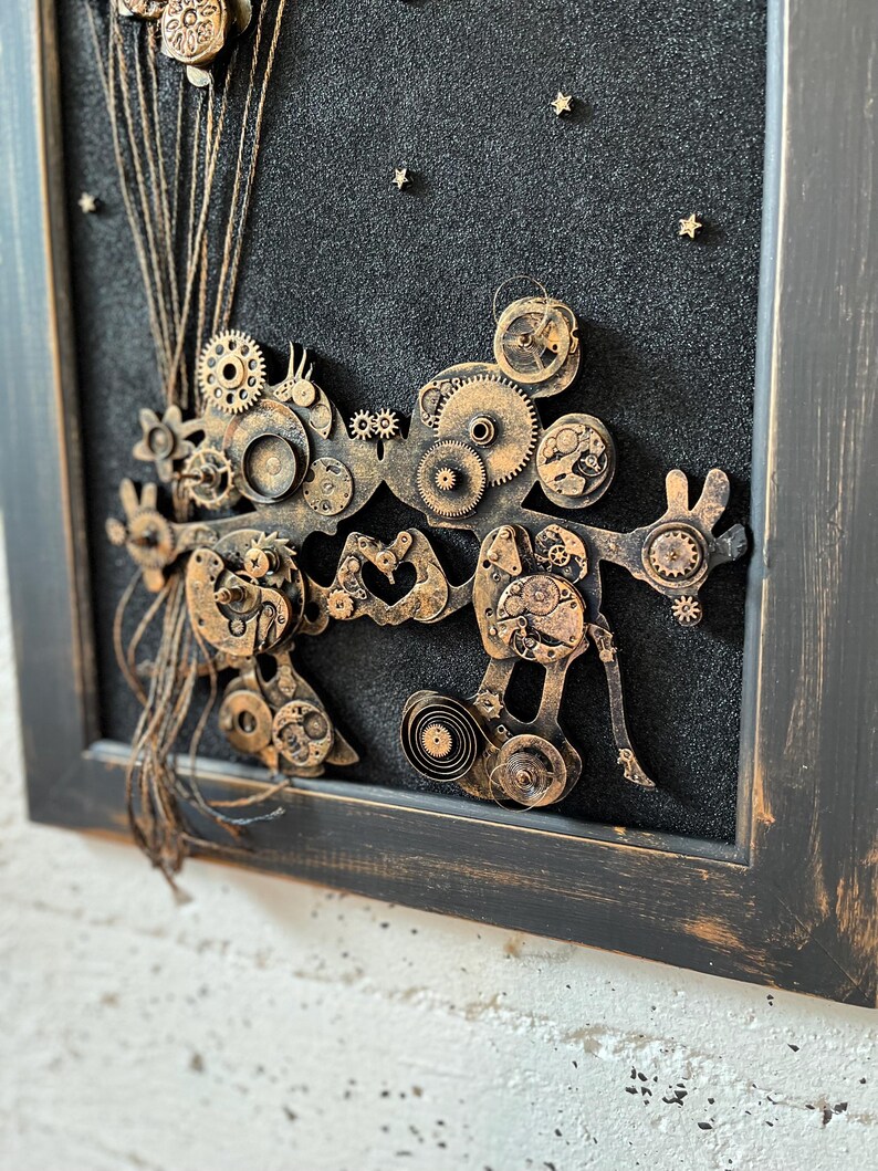 May include: A framed artwork featuring a steampunk-inspired design with gears, cogs, and other metal elements arranged in a whimsical, abstract shape. The background is a dark black with gold glitter stars.