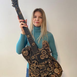 May include: A woman holds a black electric guitar with a steampunk design. The guitar is covered in gears, cogs, and other metal accents. The guitar has a gold finish and a black body.