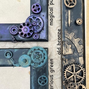 May include: Three wooden picture frames with different colored paint finishes. The frames are decorated with metal gears and have the words "mysterious green", "old bronze", and "magical purple" written on them.