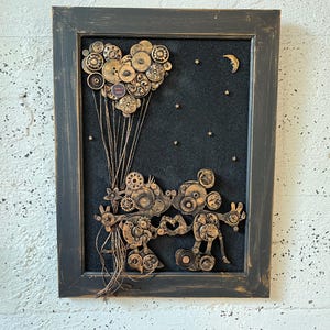 May include: A framed artwork featuring a black background with a heart-shaped arrangement of gold gears and a crescent moon. Two figures made of gold gears are holding hands, with a string of gold gears extending from their hands to the heart.
