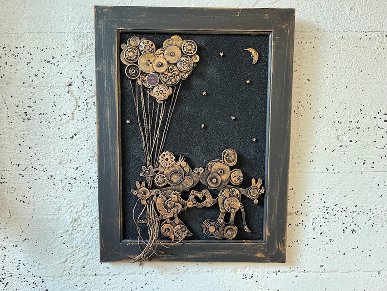 May include: A framed artwork featuring a black background with a heart-shaped arrangement of gold gears and a crescent moon. Two figures made of gold gears are holding hands, with a string of gold gears extending from their hands to the heart.
