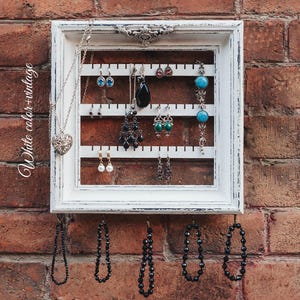 May include: A white vintage picture frame with a white background and black hooks. The frame is filled with jewellery, including necklaces, earrings, and bracelets. The jewellery is arranged in rows and columns. The frame is hanging on a brick wall.