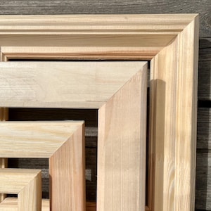 Puede incluir: Cuatro marcos de fotos de madera de diferentes tamaños, apilados uno encima del otro. Los marcos son de madera natural sin pintar, de color marrón claro.