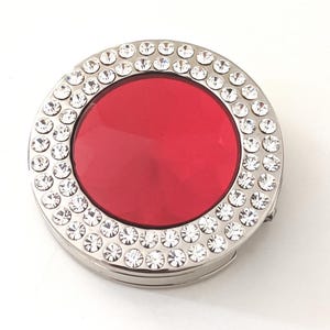 May include: A silver compact mirror with a red centre and a border of clear rhinestones.