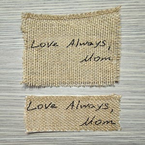 May include: Two rectangular burlap patches with frayed edges. The top patch is larger than the bottom one. Both patches have the handwritten text "Love Always, Mom" in black ink.