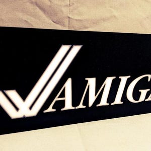 Könnte beinhalten: Ein schwarzes rechteckiges Schild mit einem weißen Logo. Das Logo ist ein stilisiertes Häkchen mit zwei Linien und dem Wort "AMIGA" in weißen Buchstaben.