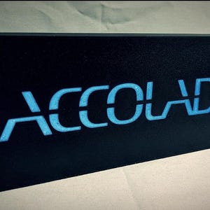 Könnte beinhalten: Ein schwarzes rechteckiges Schild mit dem Wort "ACCOLADE" in blauen Buchstaben.